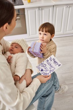 Sevimli çocuk, yeni doğan erkek kardeşi Anneler Günü 'nü elinde tutarken annesine hediye ve kartpostal veriyor.