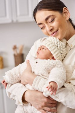Güzel, neşeli bir kadın rahat bir ev elbisesi içinde, yeni doğmuş bebeğiyle poz veriyor, modern ebeveynlik yapıyor.