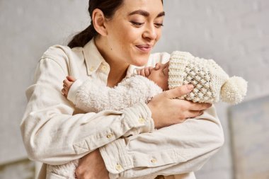 Yeni doğmuş bebeğini sevgiyle kucaklayan ev elbiseli neşeli anne, modern ebeveynlik.