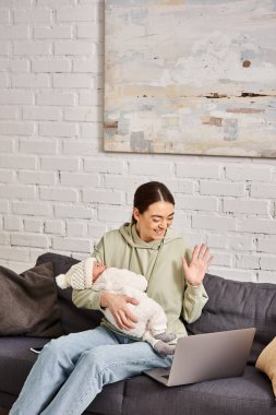 good looking joyful mother in casual attire having videocall and holding her newborn baby boy clipart