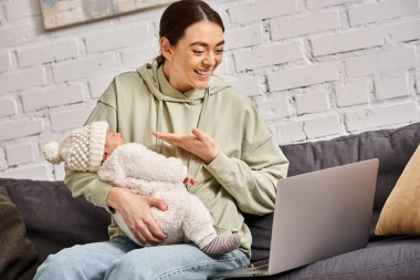 good looking joyous mother in casual attire having videocall and holding her newborn baby boy clipart