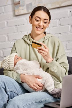 Ev elbiseleri içinde gülümseyen güzel bir anne kucağında küçük oğlunu dizüstü bilgisayarıyla elinde kredi kartıyla tutuyor.