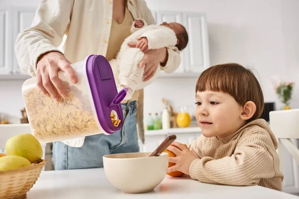 stock image joyous woman pouring corn flakes to her adorable toddler son while holding her newborn baby