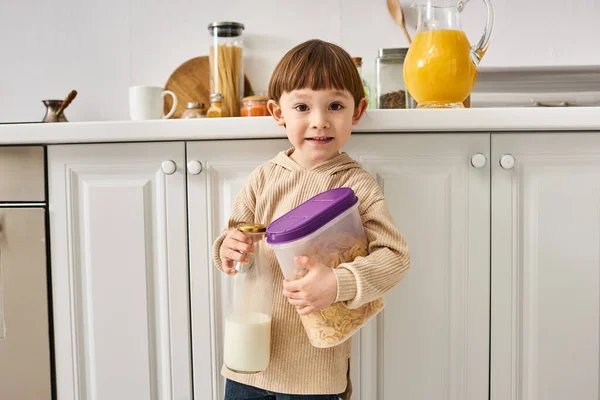 stock image adorable toddler boy in cozy homewear holding milk and corn flakes during breakfast on kitchen