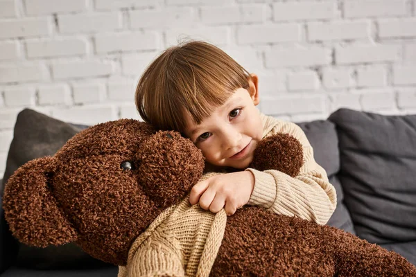 jolly little boy in homewear hugging his teddy bear while sitting on couch and looking at camera