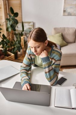 Evdeki masasında dizüstü bilgisayarı ve akıllı telefonuyla e-öğrenmeye odaklanan genç bir kız.