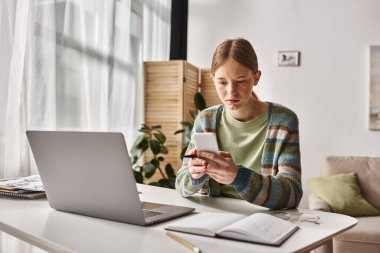 Bilgisayarın yanında otururken akıllı telefonunu kullanan konsantre genç kız, e-çalışma oturumu