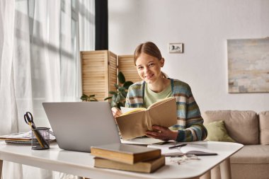 Evde e-öğrenim için dizüstü bilgisayar kullanırken gülümseyen bir kitabı olan genç kız.