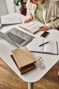 top view of book and stationery on working desk near concentrated teenage girl doing homework clipart