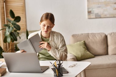 busy teenage girl holding her study book and sitting in front of a laptop at home, e-learning clipart