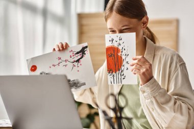 Genç kız yüzünü kapatırken sanat eserlerini gösteriyor ve bilgisayarına, resim dersine internette bakıyor.