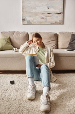 Pensive teenage girl reading her notebook and sitting near the couch with smartphone nearby clipart