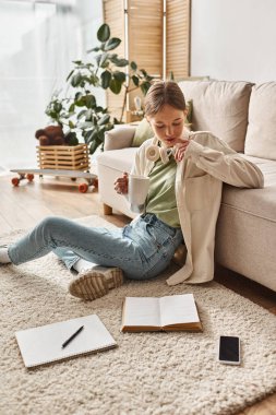 Kulaklıklı genç kız evde kahve içip halıdaki akıllı telefon ve deftere bakıyor.