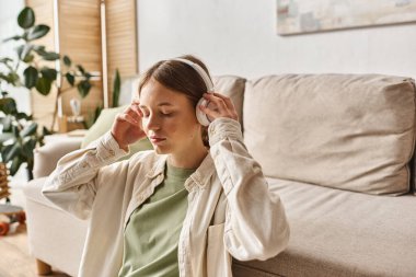 Genç kız kablosuz kulaklıklarıyla rahatlıyor ve kanepenin yanında müzik dinliyor.