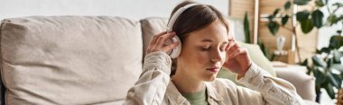 Genç Gen Z kızı kablosuz kulaklıklarıyla rahatlıyor ve kanepenin yanında müzik dinliyor.