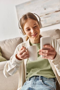 Kablosuz kulaklıklı kaygısız genç kız evde çay içerken akıllı telefon kullanıyor.