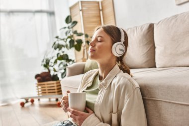 Kablosuz kulaklık takan, müzik dinleyen ve elinde çay tutan kaygısız genç kız.