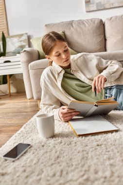 Teen girl reading book while doing homework among device, and cup of tea on carpet, generation z clipart