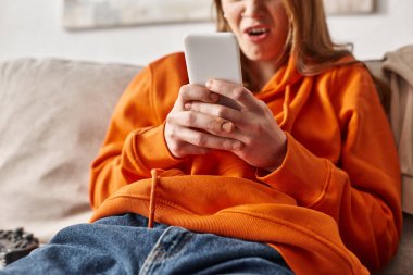 cropped and shocked teenage girl using smartphone and sitting on sofa in living room, social media clipart