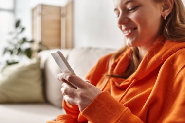Akıllı telefonunu kullanan neşeli genç kız oturma odasında kanepede oturuyor, sosyal medya kullanıcısı