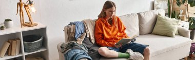 teenage girl reading book while sitting on messy sofa next to pile of clothes in apartment, banner clipart