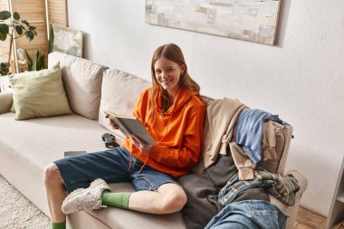 happy teenager reading book while sitting on sofa next to messy pile of clothes in apartment clipart