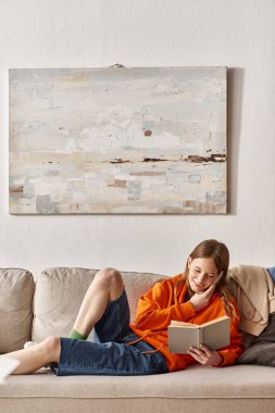 teenager girl holding book during phone call and sitting on sofa next to messy pile of clothes clipart