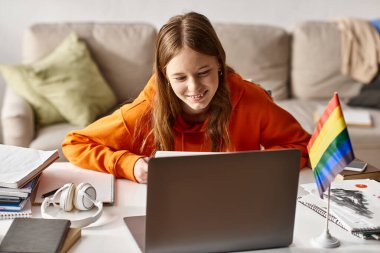 Joyful teenage girl looking at laptop and enjoying e-learning next to headphones and pride flag clipart