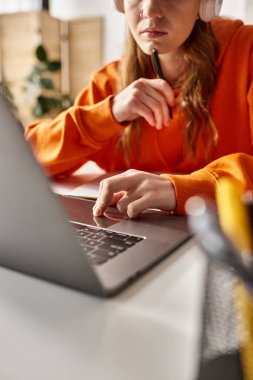 Kablosuz kulaklık takan genç bir kızın evde bilgisayarla internetten e-öğrenme görüntüsü.