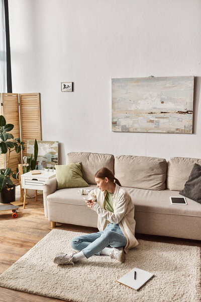 счастливая девочка-подросток, используя свой смартфон, сидя на ковре рядом с диваном, цифровые устройства