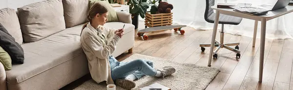 年轻的少女坐在客厅沙发边的地毯上 一边用智能手机 — 图库照片