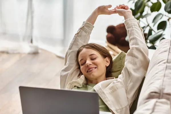 stock image cheerful teenage girl lying with her laptop and stretching on comfortable couch, distance learning