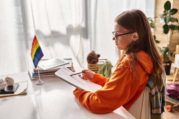pensive teenage girl in glasses drawing a sketch, immersed in creative process near pride flag