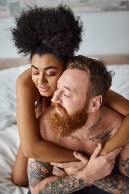 Seksi, çeşitli, kıvırcık Afrikalı Amerikalı kadın dövmeli erkek arkadaşını yatak odasında kucaklıyor.