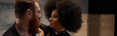 Kıvırcık saçlı Afro-Amerikalı kadın. Sakallı, afiş takan dövmeli erkek arkadaşını baştan çıkarıyor.