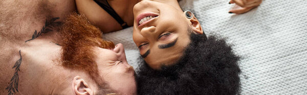 Intimate multicultural couple laughing and holding hands on bed in light-filled bedroom, banner