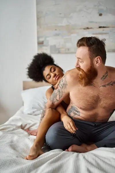 sexy diverse couple, african american woman embracing her tattooed man on a bed in a bright room