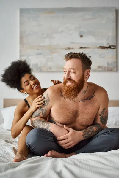 Joyful diverse couple, african american woman embracing her tattooed man on a bed in a bright room