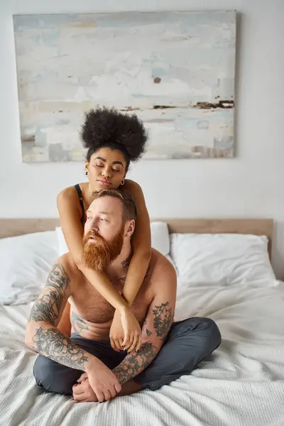 diverse couple, african american woman embracing her bearded tattooed man on a bed in a bright room