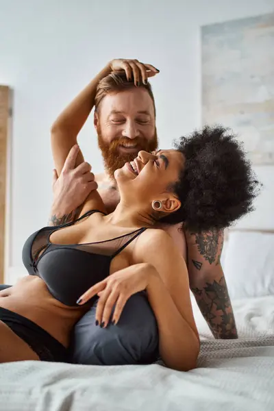 bearded man with tattoos laughing with african american girlfriend in lingerie, diverse couple