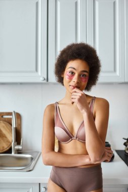 Modern mutfakta duran, gözlerinin altında pembe bantlı, sütyenli Afro-Amerikan kadını cezbediyor.