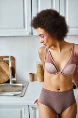 Modern mutfakta, gözlerinin altında pembe yamalar olan, sütyen takmış neşeli Afro-Amerikalı kadın.