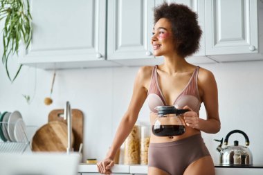 Güneş ışığıyla aydınlanmış modern bir mutfakta elinde kahve fincanı tutan, göz bantları olan neşeli, Afro-Amerikalı bir kadın.