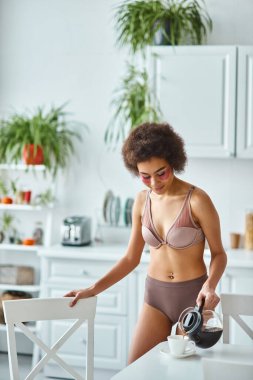 Göz bantlı Afro-Amerikalı kadın mutfaktaki porselen bardağa kahve dolduruyor.