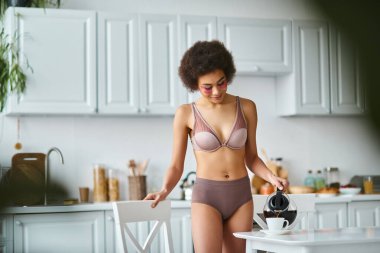 Kıvırcık, mutlu, göz bantlı Amerikalı kadın mutfaktaki porselen bardağa kahve dolduruyor.