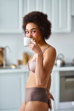İç çamaşırlı, sabah kahvesini yudumlayan, göz bantlı mutlu Afrikalı Amerikalı kadın.