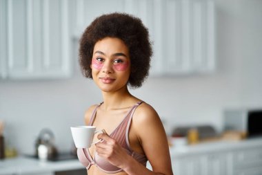 Güzel Afro-Amerikalı kadın iç çamaşırlı ve fincanda kahvenin tadını çıkarıyor.
