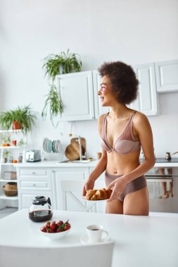 İç çamaşırlı mutlu Afro-Amerikan kadın kahvaltıda kruvasan ve çilek servis ediyor.