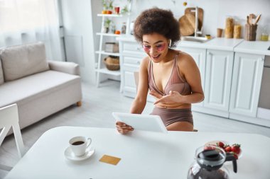 Mutlu Afro-Amerikan kadın iç çamaşırı ve göz bantları ile modern mutfakta görüntülü sohbet yapıyor.