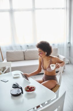 Mutlu Afro-Amerikan kadın iç çamaşırlarıyla tablet kullanıyor ve sabahları kahve içiyor.